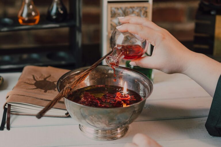 Home Close-up of a hand pouring red potion into a flame-lit bowl, suggesting a magical ritual.